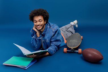 Genç bir adam okuyor, etrafı kaykayla çevrili ve mavi bir zemine karşı futbol oynuyor..