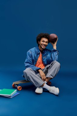 Genç, şık bir adam parlak bir stüdyoda elinde futbol topu tutarken kaykayla rahatlıyor..