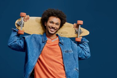 Kıvırcık saçlı neşeli bir genç adam kaykayını omuzlarının üzerinde tutarken gülümsüyor..