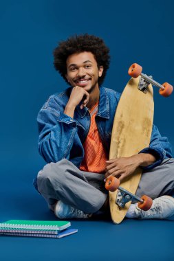 Genç bir adam parlak mavi bir yüzeyde, elinde kaykayla rahatça gülümsüyor..