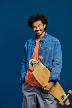 Kıvırcık saçlı neşeli genç bir adam mavi bir duvarın yanında kaykayla poz veriyor..