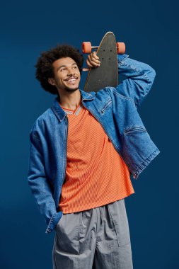 Genç adam kaykayını parlak mavi bir arka plana karşı gururla gösteriyor..