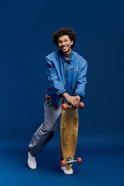 Neşeli genç bir adam mavi bir stüdyoda kot ceketli bir kaykayı güvenle tutuyor..