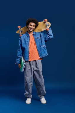 Genç bir adam parlak mavi bir arka plana karşı bir kaykay ve kitap tutuyor..