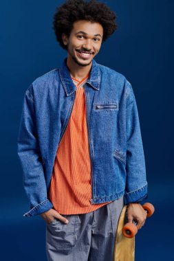 Parlak elbiseli genç adam bir kaykayla gülümsüyor, stüdyoda neşe saçıyor..