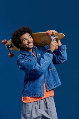 Kıvırcık saçlı genç bir adam gülümsüyor, elinde mavi bir arka planda kaykay tutuyor..