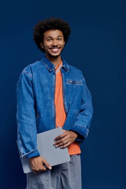 Gülümseyen genç adam dizüstü bilgisayarla poz veriyor, mavi arka planda şık bir kıyafet sergiliyor..