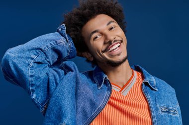 Kıvırcık saçlı, turuncu çizgili gömlek ve kot ceketli genç bir adam gülümsüyor..