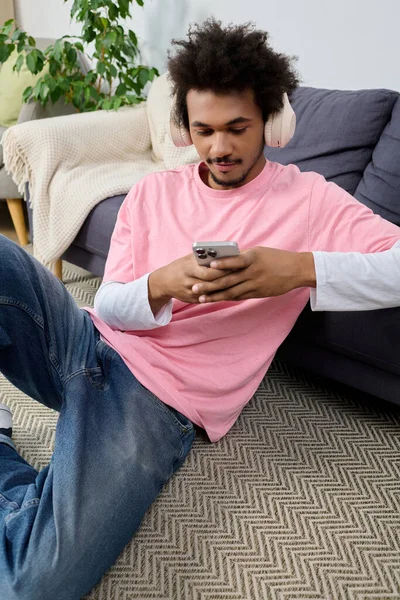 Pembe gömlekli genç adam yerde oturuyor, akıllı telefonunu kullanıyor ve müzik dinliyor..