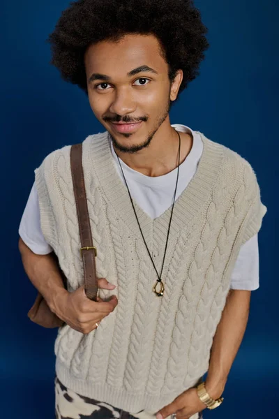 Rahat bir yelek giymiş kendine güvenen genç bir adam mavi arka planda bir çantayla poz veriyor..