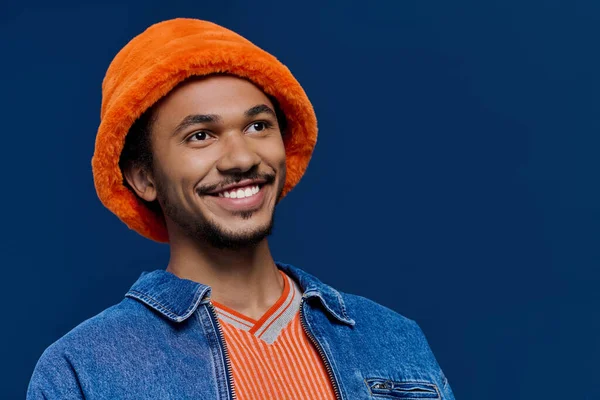 Kot ceketli ve turuncu şapkalı neşeli bir genç sıcak bir gülümsemeyle duruyor..