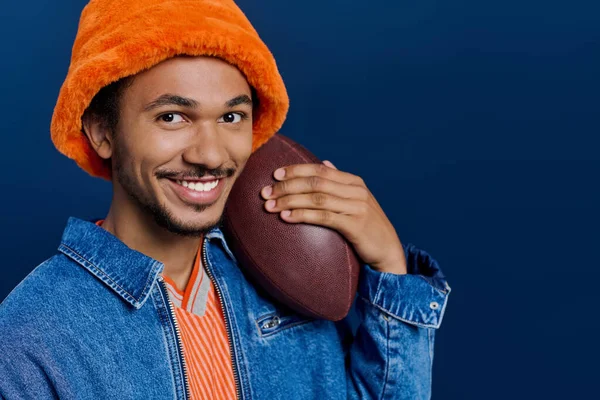 Turuncu şapkalı neşeli bir genç adam elinde bir futbol topuyla büyüleyici bir poz veriyor..