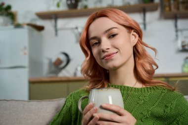 Güzel genç bir kadın, şık ve rahat bir yaşam alanında sıcak bir kupayı tutarken gülümsüyor..