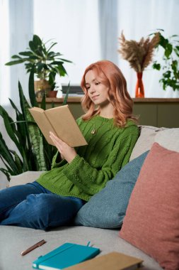 A beautiful young woman enjoys her time reading on a comfortable sofa surrounded by greenery.