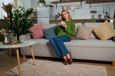 Genç bir kadın, elinde bir kupayla, yumuşak yastıklarla huzurlu bir anın tadını çıkarıyor..