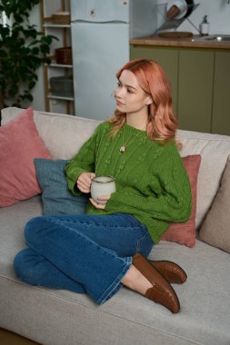 Genç bir kadın rahat bir koltukta oturur, çayını yudumlar ve evde huzurlu bir anın tadını çıkarır..