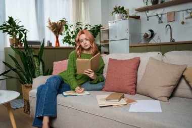 A beautiful young woman sits on a sofa, writing in her notebook while surrounded by greenery.