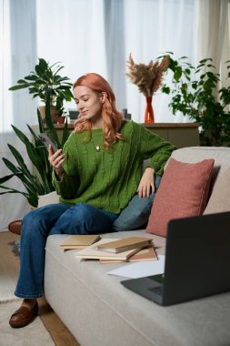 Relaxing on a couch, a beautiful young woman engages with her phone in a green sweater.