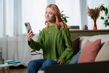 A young woman in a vibrant green sweater smiles as she interacts with her smartphone at home.