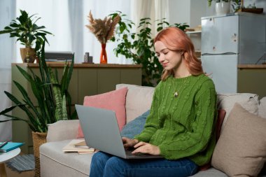 A young woman relaxes on a cozy sofa, focused on her laptop amid vibrant decor.