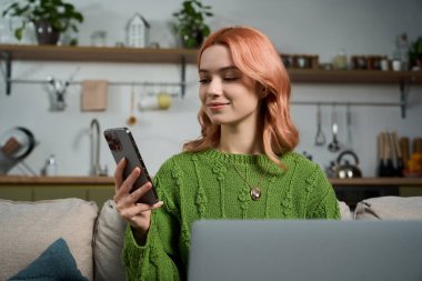 Pembe saçlı genç bir kadın rahat bir ortamda telefonunu kullanırken gülümsüyor..