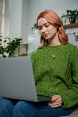 A young woman with beautiful pink hair is focused on her laptop, surrounded by plants.