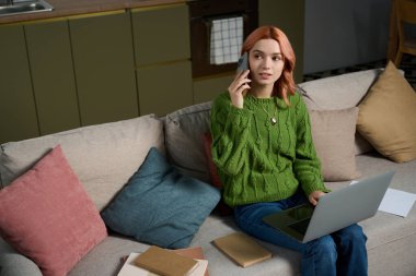 Genç bir kadın kanepede dinlenirken telefonuyla sohbet ediyor ve bilgisayarını kullanıyor..