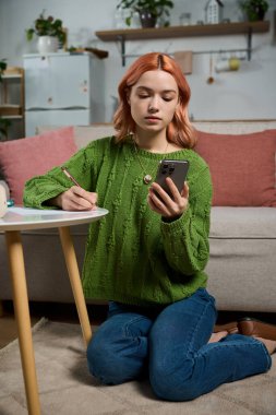 Beautiful young woman sits on the floor, taking notes while checking her phone in a stylish room.