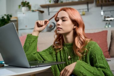 Yeşil kazaklı genç bir kadın sıcak, davetkar bir mekanda dizüstü bilgisayarına dikkatle bakar..