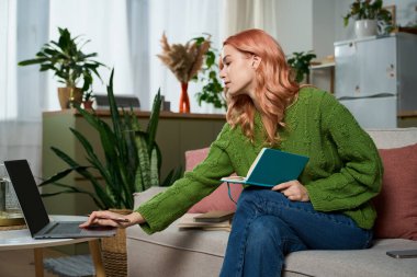 A young woman with pink hair sits on a couch, using her laptop and holding a notebook.