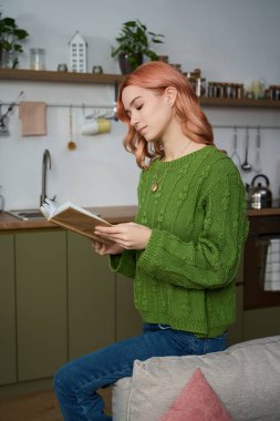 A young woman reads intently in a warm, inviting kitchen filled with cozy details.