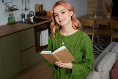 Sıcak ve davetkar bir mutfakta elinde kitap tutarken gülümseyen pembe saçlı güzel bir genç kadın..