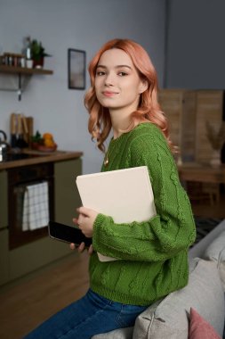 A cheerful young woman with pink hair holds a notebook and phone in a stylish kitchen.