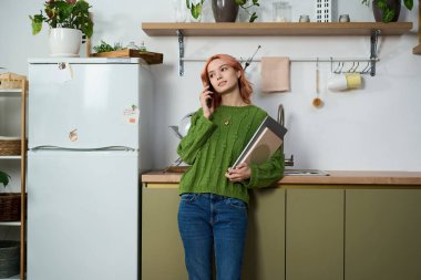 Parlak ışıklandırılmış mutfak sahnesi. Telefonda konuşan yeşil kazaklı genç bir kadın..