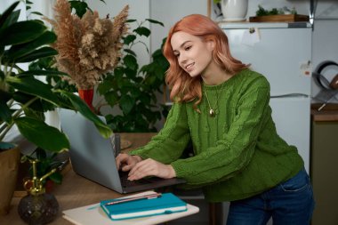 Saçları dalgalı genç bir kadın sıcak, yeşil bir ortamda dizüstü bilgisayarına odaklanır..