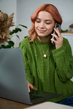 Güzel genç bir kadın bilgisayarında yazarken telefonuyla konuşuyor. Etrafı bitkilerle çevrili..