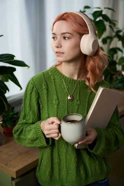 Yeşil kazaklı şık bir kadın müzik dinlerken elinde bir fincan kahve tutuyor..