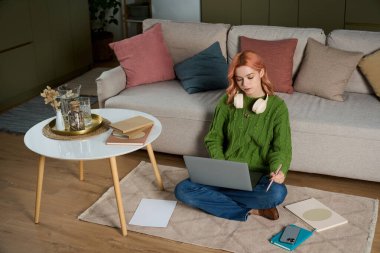 Odaklanmış genç bir kadın şık bir oturma odasında rahatça otururken dizüstü bilgisayarıyla çalışıyor..