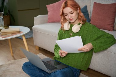A beautiful young woman sits comfortably on the floor, reading papers with a laptop beside her.