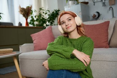 Güzel genç bir kadın canlı yeşil bir kazakla rahatça otururken müzik dinler..