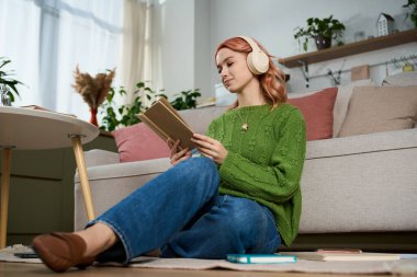 Güzel genç bir kadın modern evinde bir kitap ve müzikle huzurlu bir anın tadını çıkarıyor..