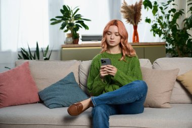 Pembe saçlı genç bir kadın rahat bir şekilde koltukta oturuyor, telefonuna dalmış, dinleniyor..