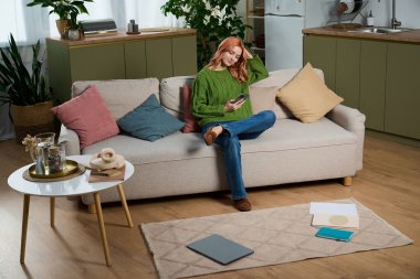 Güzel, genç bir kadın şık bir oturma odasındaki kanepede rahat bir şekilde oturuyor, telefonunu kaydırıyor..