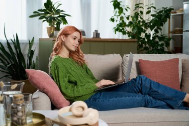 A beautiful young woman with pink hair relaxes on a sofa, working on her laptop in a cozy setting.