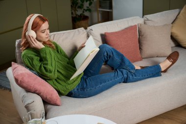 Yeşil kazaklı genç bir kadın kanepede rahatça oturur, müzik dinler ve kitap okur..