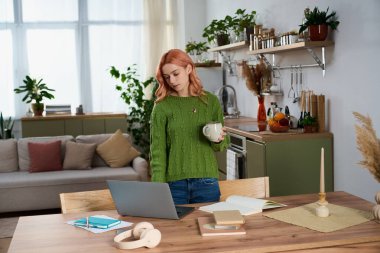 Pembe saçlı genç bir kadın sıcak bir mutfakta duruyor, çayını yudumluyor ve gününü yaşıyor..