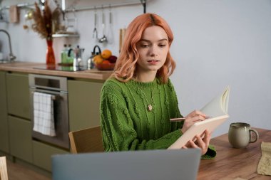 In a stylish kitchen, a young woman with vibrant hair sits at a table, focused on her notebook.