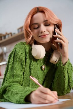 Güzel, genç bir kadın, fikirlerini şık bir masada yazarken telefonla konuşmaktan zevk alır..