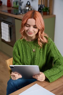 A cheerful young woman in a green sweater sits in a lovely kitchen, focused on her tablet.