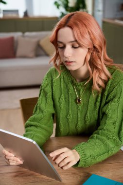 A beautiful young woman with pastel hair intently uses her tablet at a wooden table.